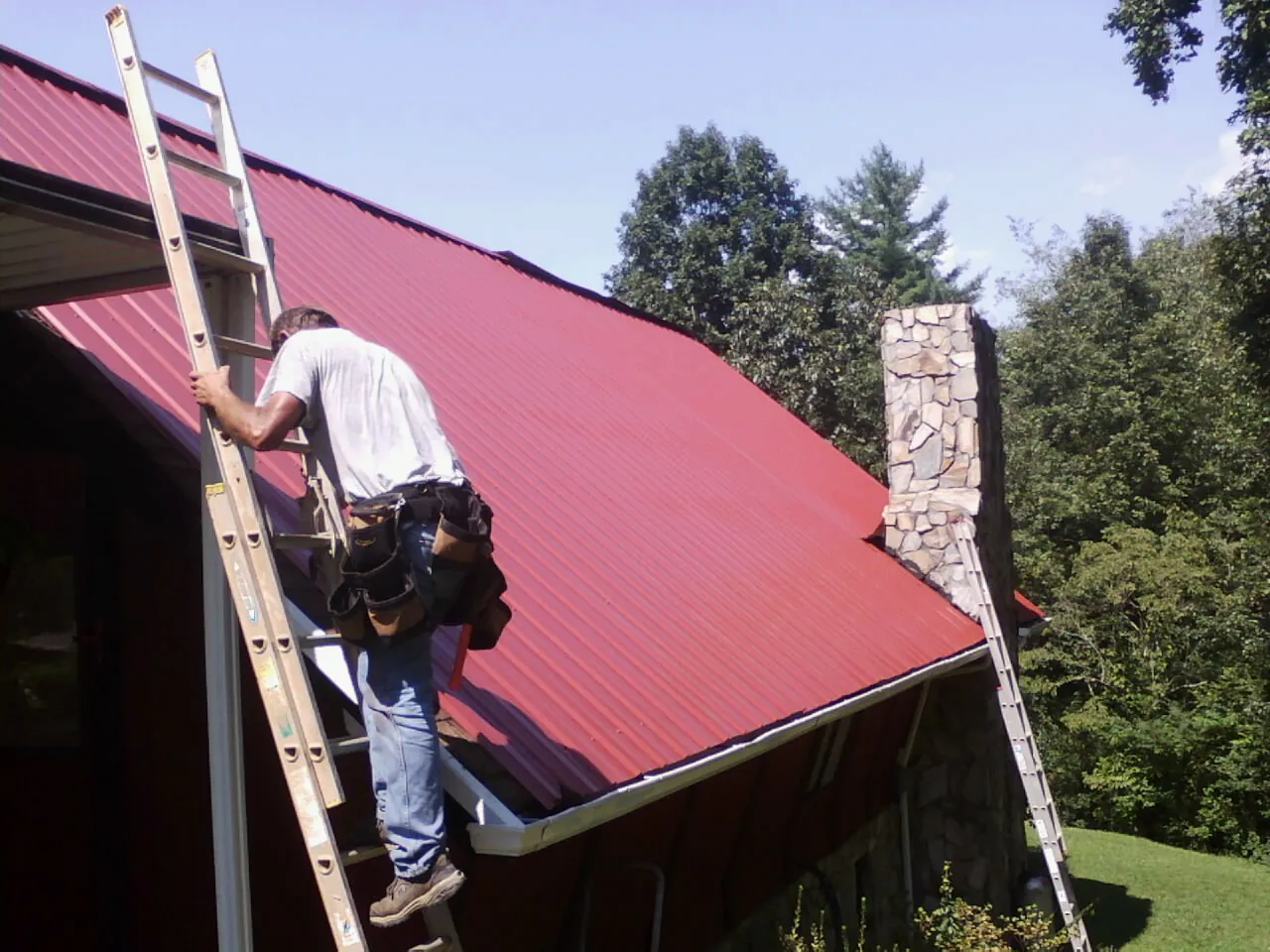 Roofer on a ladder beside a red metal roof and stone chimney — Soffit & Fascia Repair by Roofing Brothers in Kamiah